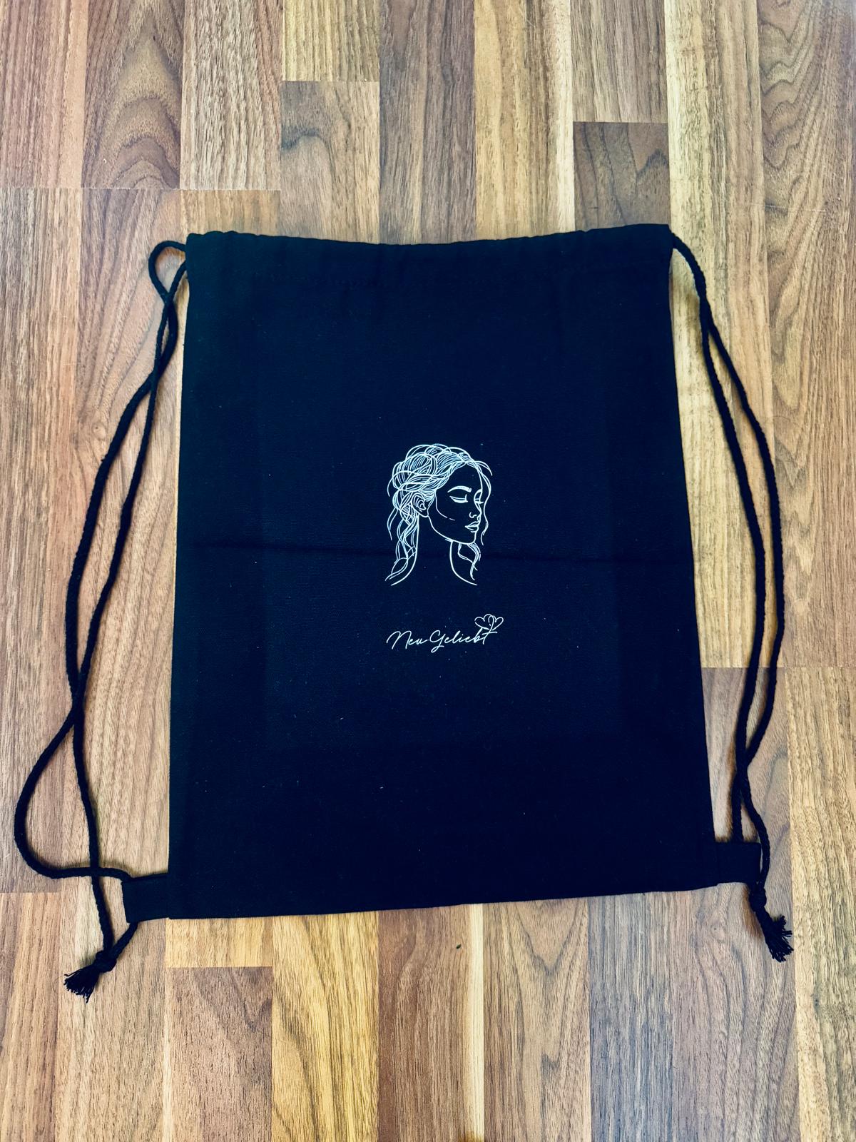 Schwarzer Rucksack aus Canvas mit Kordelzug und NeuGeliebt Logo auf dem Parkettboden.