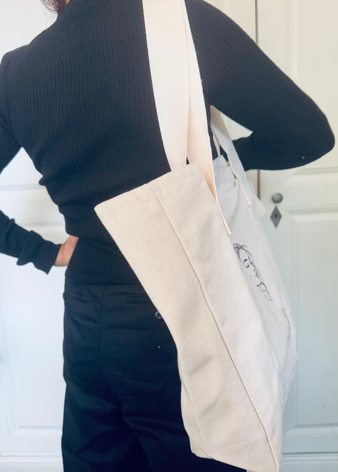 Eine Frau den Rücken zugewandt, mit Beutel aus naturfarbenen Canvas mit Frauenkopf und Logo von NeuGeliebt.